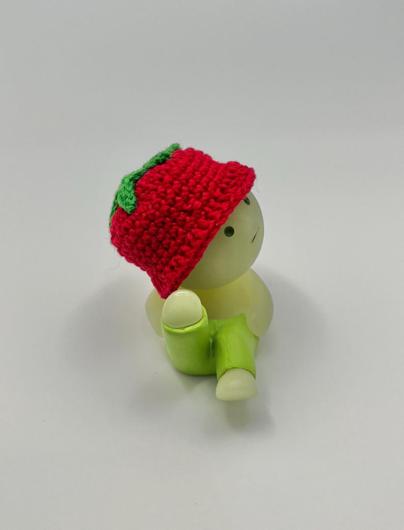 May include: A small, white figurine wearing a red crocheted hat with a green leaf detail and green pants. The figurine is sitting with its legs crossed.