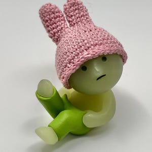 May include: A small, green, glow-in-the-dark figurine wearing a pink crocheted bunny hat and green pants. The figurine has a sad expression on its face.