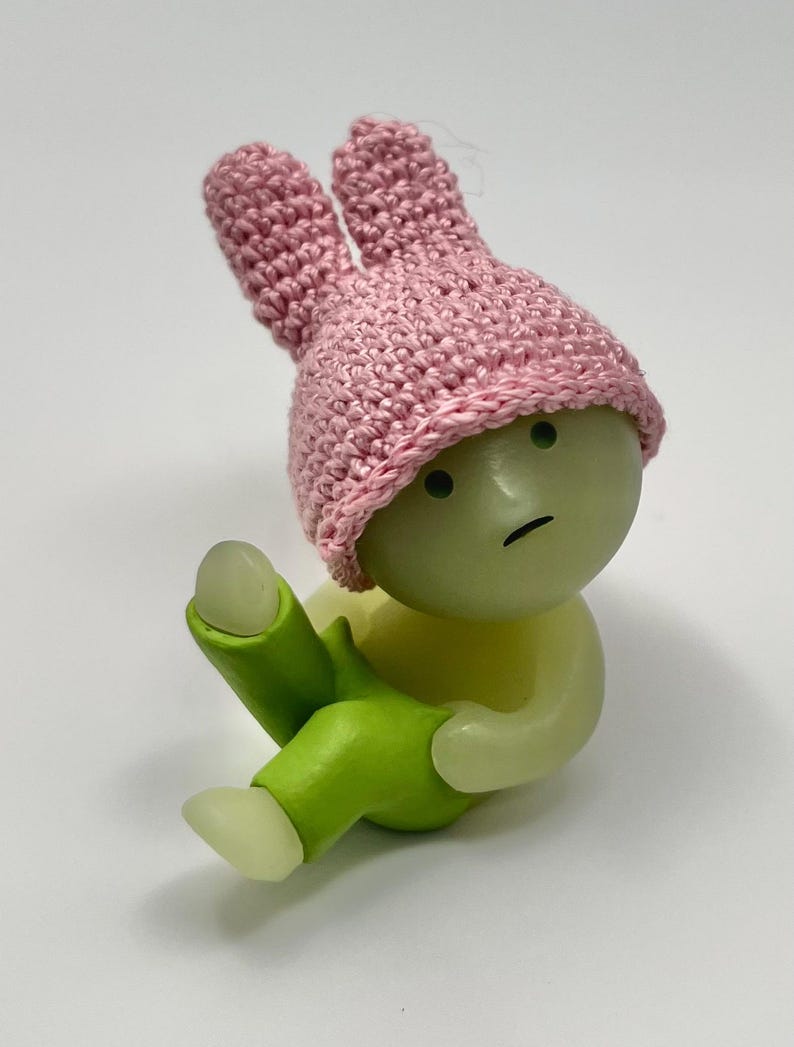 May include: A small, green, glow-in-the-dark figurine wearing a pink crocheted bunny hat and green pants. The figurine has a sad expression on its face.