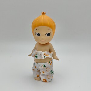 May include: A small, orange-haired doll wearing a white and orange floral print outfit. The doll has a round head and large, black eyes.