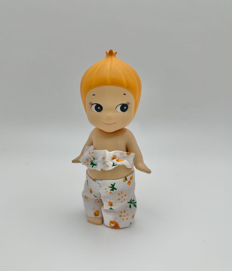 May include: A small, orange-haired doll wearing a white and orange floral print outfit. The doll has a round head and large, black eyes.
