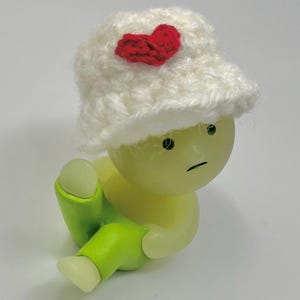 May include: A small, green, plastic figurine wearing a white crocheted hat with a red heart on top. The figurine has a sad expression and is sitting with its legs crossed.