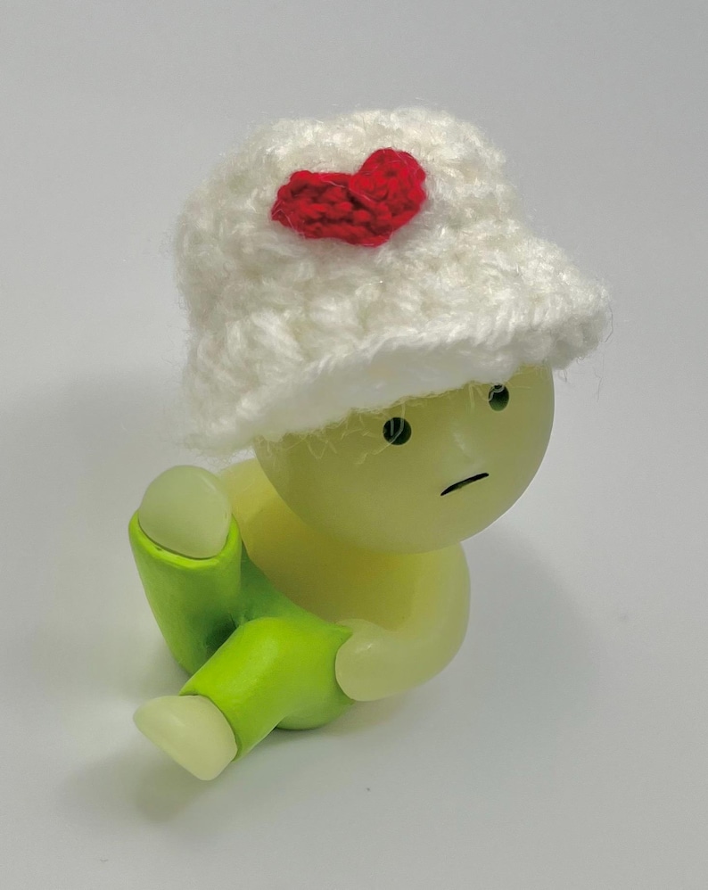 May include: A small, green, plastic figurine wearing a white crocheted hat with a red heart on top. The figurine has a sad expression and is sitting with its legs crossed.
