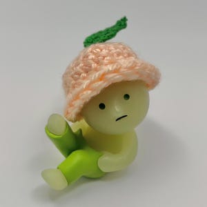 May include: A small, green and peach-colored figurine with a sad expression. The figurine is wearing a pink crocheted hat with a green leaf on top. The figurine is sitting with its legs crossed.