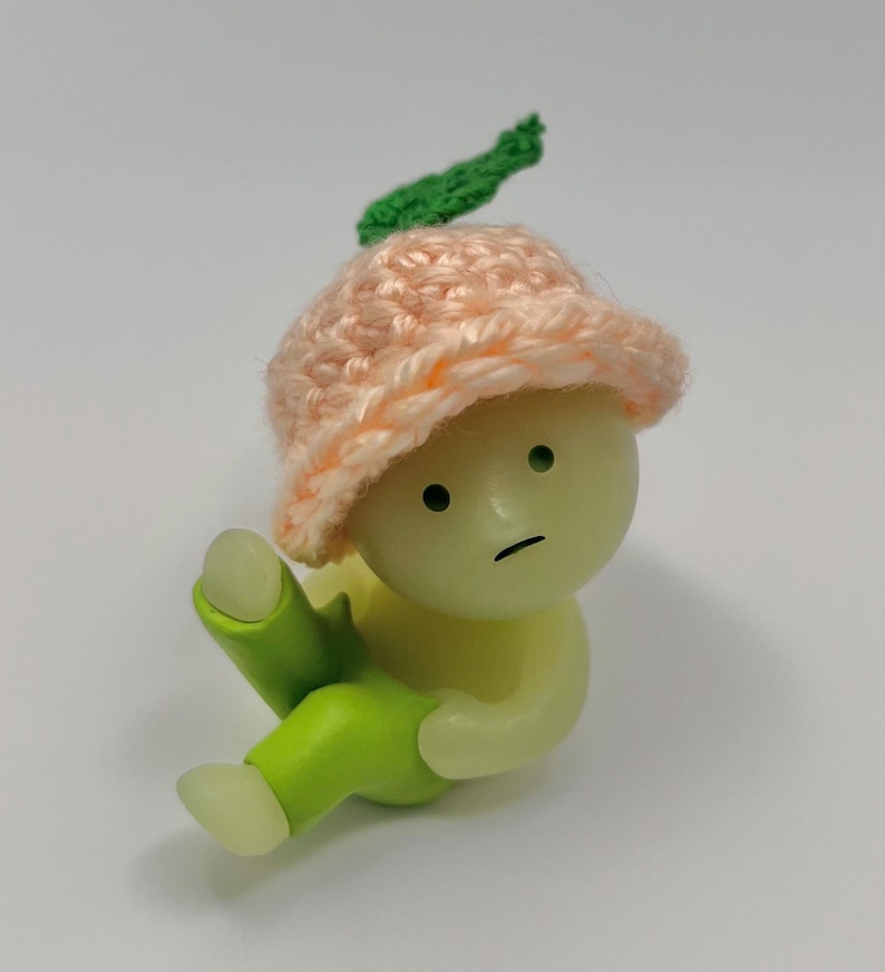 May include: A small, green and peach-colored figurine with a sad expression. The figurine is wearing a pink crocheted hat with a green leaf on top. The figurine is sitting with its legs crossed.