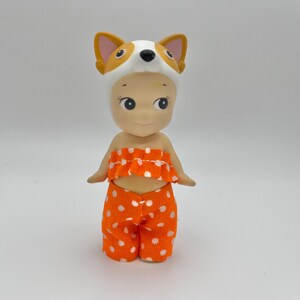 May include: A small doll wearing an orange and white polka dot outfit and a white fox hat.