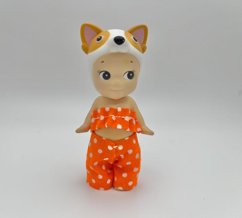 May include: A small doll wearing an orange and white polka dot outfit and a white fox hat.