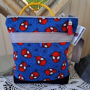 May include: A blue tote bag with a Spider-Man face and spiderweb pattern. The bag has a grey band, a black base, and a red tassel zip pull. It has a yellow handle and a tag that reads "Elvira's Handmade Corner."