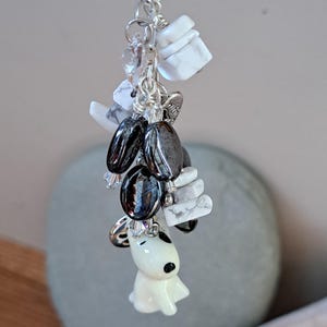 May include: A white ceramic dog charm with black eyes and nose, hanging from a silver chain with black and white beads, white stones, and a crystal bead.