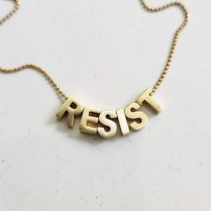 Peut inclure: Un collier doré avec le mot "RESIST" épelé en lettres majuscules. Les lettres sont reliées par une chaîne délicate. Le collier est présenté sur une surface blanche.