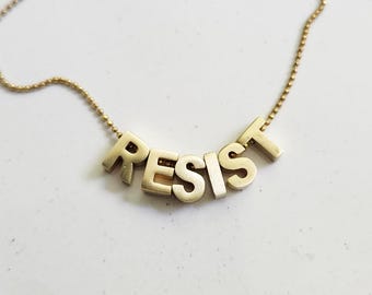 May include: A gold-colored necklace with the word "RESIST" spelled out in block letters. The letters are connected by a delicate chain. The necklace is displayed on a white surface.