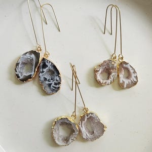 May include: Three pairs of dangle earrings featuring natural geode stones with gold-colored metal accents. The geodes display shades of gray, white, and brown. The earrings are displayed on a white surface.