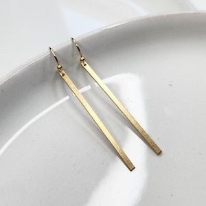 May include: A pair of gold-toned metal drop earrings featuring long, slender, rectangular bar pendants suspended from simple curved ear wires. The metal surface has a subtle brushed texture, and the design is clean, linear, and symmetrical.