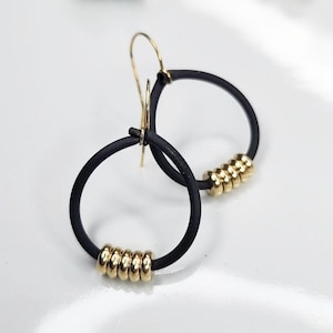 May include: A pair of black hoop earrings with gold-colored accents. Each earring features a circular black band with a series of small, gold-colored beads. The earrings have a simple, modern design.