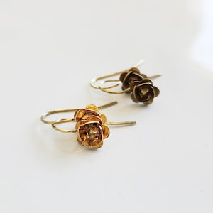 May include: A pair of gold-colored earrings featuring a rose design. The earrings have a hook closure and are set against a white background. The rose petals are detailed, and the earrings appear to be lightweight.