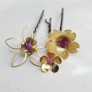 May include: Three gold-toned hair pins featuring floral designs. One pin has a wire-outline flower with a purple bead center. Another pin features a textured, glittery gold flower with a pink bead center. A third small, smooth gold flower pin has a pink bead.