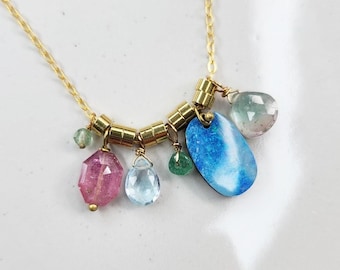 Puede incluir: Un collar de cadena dorada con una serie de pequeñas cuentas cilíndricas de oro y múltiples dijes de piedras preciosas colgantes, que incluyen una piedra rosa facetada, una gota azul claro, una cuenta verde pequeña, una piedra azul ovalada grande y una gema verde pálido.