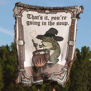 Pode incluir: Uma manta tecida com um design humor&iacute;stico. A manta apresenta um sapo de desenho animado com um chap&eacute;u de bruxa, mexendo numa panela sobre uma fogueira. O texto diz "That's it, you're going in the soup". A borda tem um padr&atilde;o decorativo.