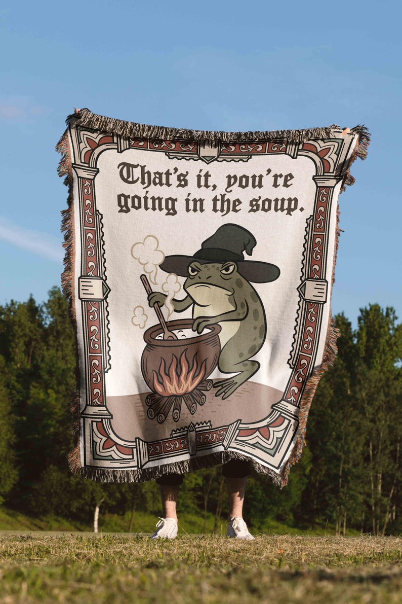 Pode incluir: Uma manta tecida com um design humor&iacute;stico. A manta apresenta um sapo de desenho animado com um chap&eacute;u de bruxa, mexendo numa panela sobre uma fogueira. O texto diz "That's it, you're going in the soup". A borda tem um padr&atilde;o decorativo.