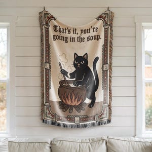 Puede incluir: Manta tejida con un gato negro removiendo un caldero sobre un fuego. El texto "That's it, you're going in the soup" se muestra en la parte superior. La manta tiene un fondo crema con bordes decorativos y flecos.