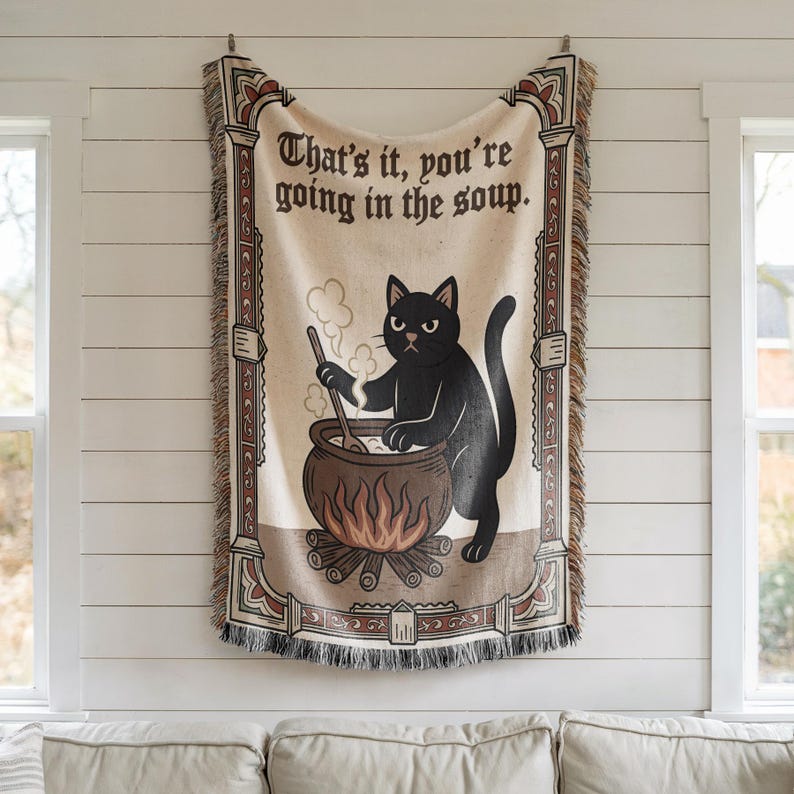 Puede incluir: Manta tejida con un gato negro removiendo un caldero sobre un fuego. El texto "That's it, you're going in the soup" se muestra en la parte superior. La manta tiene un fondo crema con bordes decorativos y flecos.