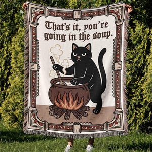 Puede incluir: Una manta crema con un borde decorativo y el texto "That's it, you're going in the soup." Un gato negro revuelve un caldero sobre un fuego. La manta tiene un borde con flecos.