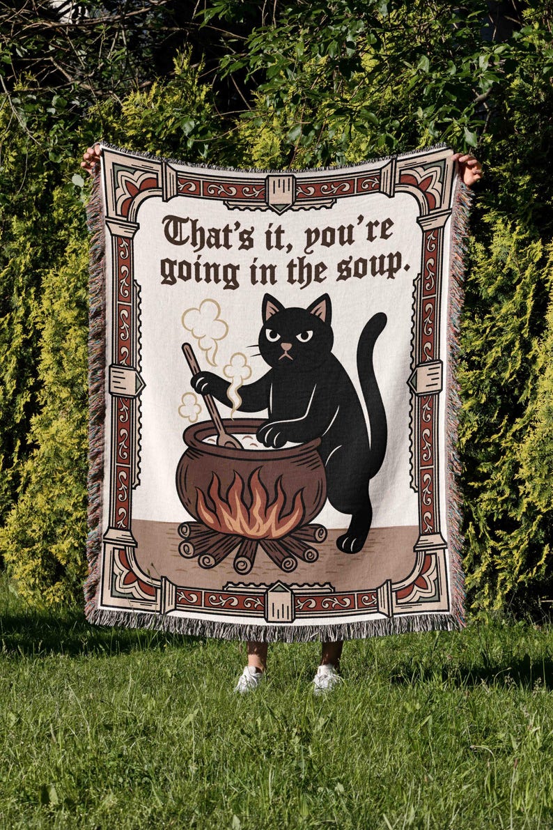 Puede incluir: Una manta crema con un borde decorativo y el texto "That's it, you're going in the soup." Un gato negro revuelve un caldero sobre un fuego. La manta tiene un borde con flecos.