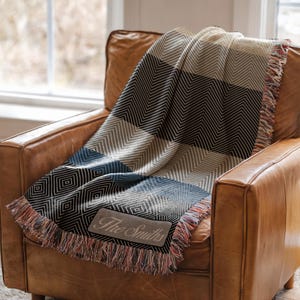May include: A woven throw blanket with a geometric pattern in shades of black, gray, and cream, draped over a brown leather armchair. The blanket has a fringed edge and a rectangular patch that reads "The Smiths".