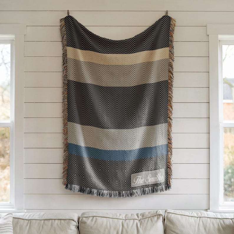 May include: A woven blanket with a striped pattern in shades of black, beige, gray, and blue, with a fringed edge. The blanket is hanging on a white wall. The text "The Smiths" is visible on a rectangular patch.