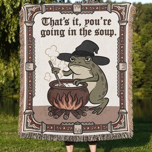 Pode incluir: Um cobertor retangular com uma ilustra&ccedil;&atilde;o de um sapo usando um chap&eacute;u de bruxa, mexendo em um caldeir&atilde;o sobre uma fogueira. O texto "That's it, you're going in the soup" &eacute; exibido no topo. O cobertor tem uma borda com franjas e uma moldura decorativa.