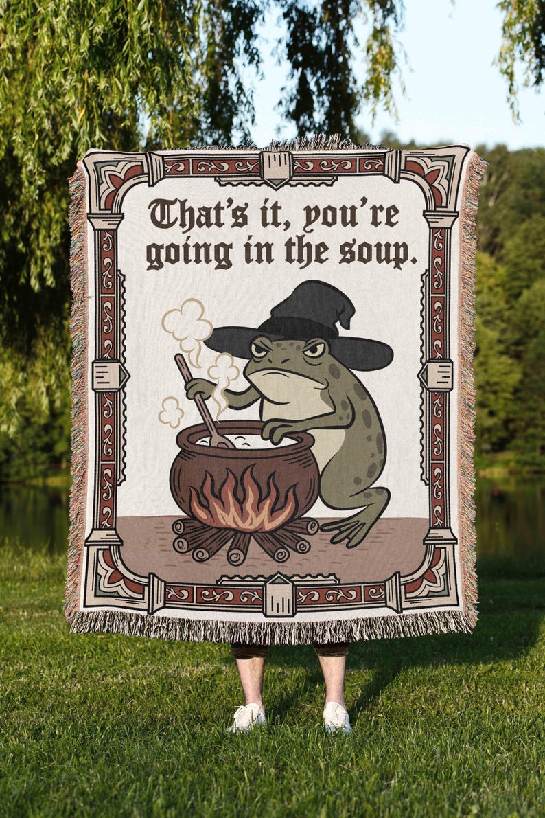 Pode incluir: Um cobertor retangular com uma ilustra&ccedil;&atilde;o de um sapo usando um chap&eacute;u de bruxa, mexendo em um caldeir&atilde;o sobre uma fogueira. O texto "That's it, you're going in the soup" &eacute; exibido no topo. O cobertor tem uma borda com franjas e uma moldura decorativa.