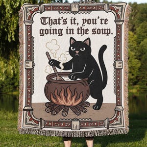 Puede incluir: Una manta tejida con un gato negro removiendo un caldero sobre un fuego, con el texto "That's it, you're going in the soup." La manta tiene un borde decorativo en tonos marr&oacute;n y crema, con flecos.