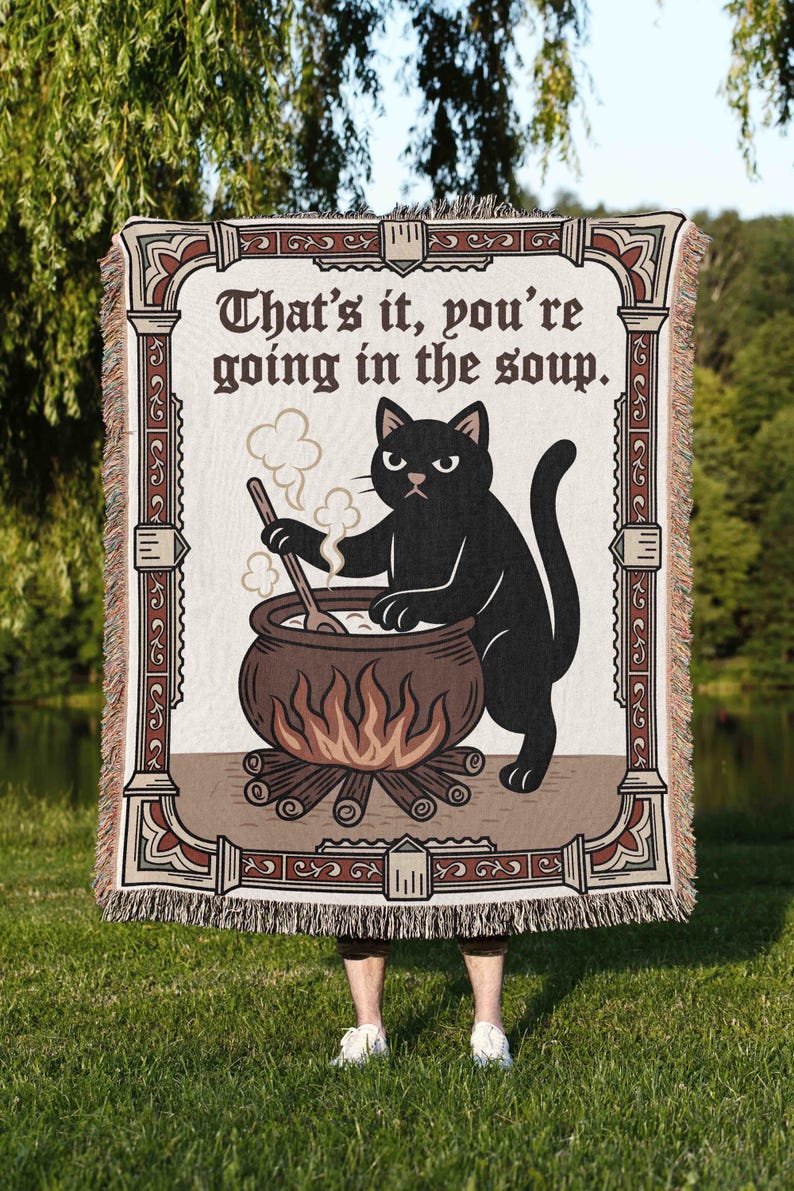 Puede incluir: Una manta tejida con un gato negro removiendo un caldero sobre un fuego, con el texto "That's it, you're going in the soup." La manta tiene un borde decorativo en tonos marr&oacute;n y crema, con flecos.