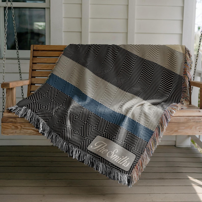 May include: A woven throw blanket with a geometric pattern in black, tan, and blue, draped over a wooden swing. The blanket features a diamond pattern and a rectangular patch with the text "The Smiths". The blanket has fringed edges.
