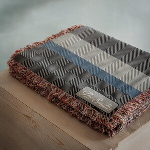 May include: A folded throw blanket with a geometric pattern in shades of gray, blue, and white. The blanket has a fringed edge in red, orange, and brown. A rectangular patch with the text "The Smiths" is sewn on the blanket.