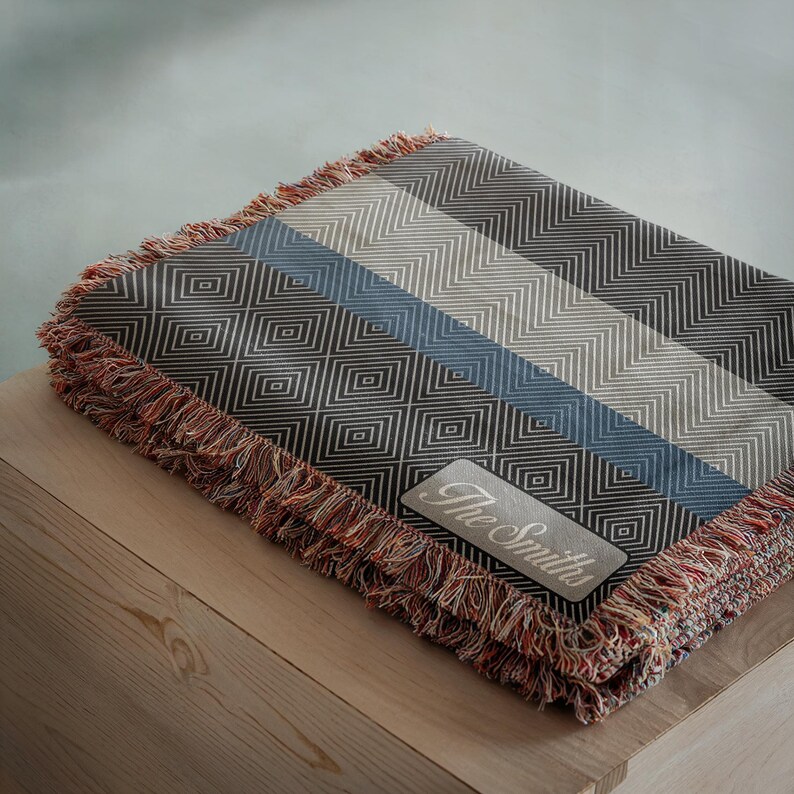 May include: A folded throw blanket with a geometric pattern in shades of gray, blue, and white. The blanket has a fringed edge in red, orange, and brown. A rectangular patch with the text "The Smiths" is sewn on the blanket.
