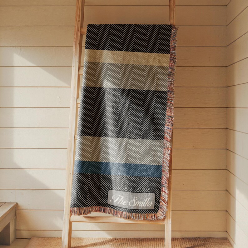 May include: A woven throw blanket with a striped pattern in shades of black, gray, beige, and blue, draped over a wooden ladder. The blanket has a fringed edge and a rectangular patch with the text "The Smiths".