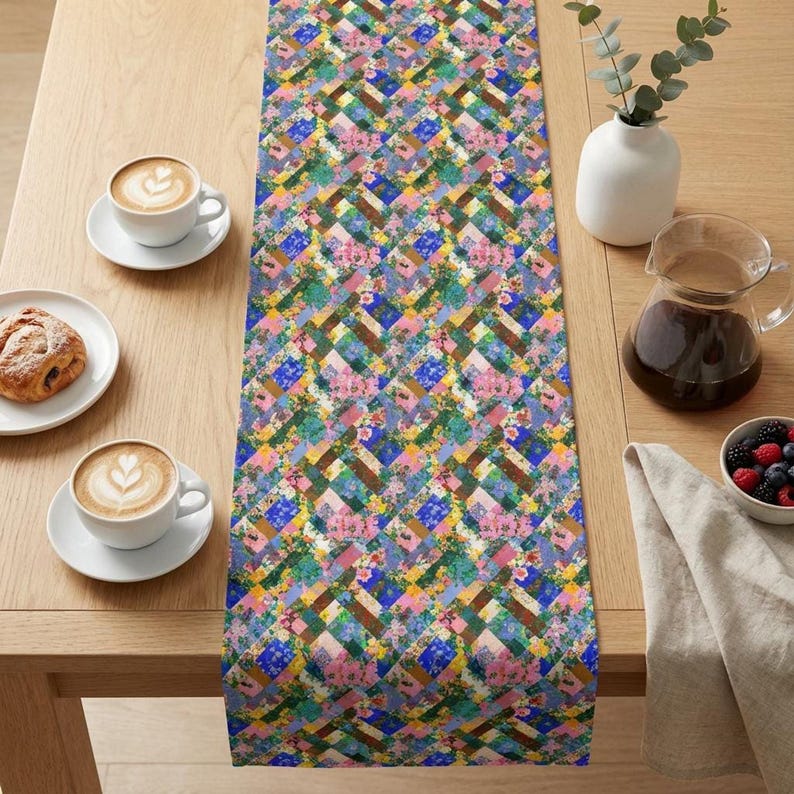 A long fabric table runner featuring a vibrant patchwork pattern of multicolored floral squares, pink blossoms, and woven blue and green blocks. The runner is styled down the center of a light oak wood dining table set with two latte coffee cups, a pastry on a plate, a glass coffee carafe, and a small bowl of fresh berries.
