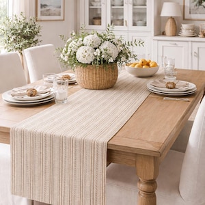 May include: A dining room table set for a meal. The wooden table is adorned with a woven table runner, white plates, and a basket of white flowers. Overhead, two woven pendant lights hang from the ceiling.