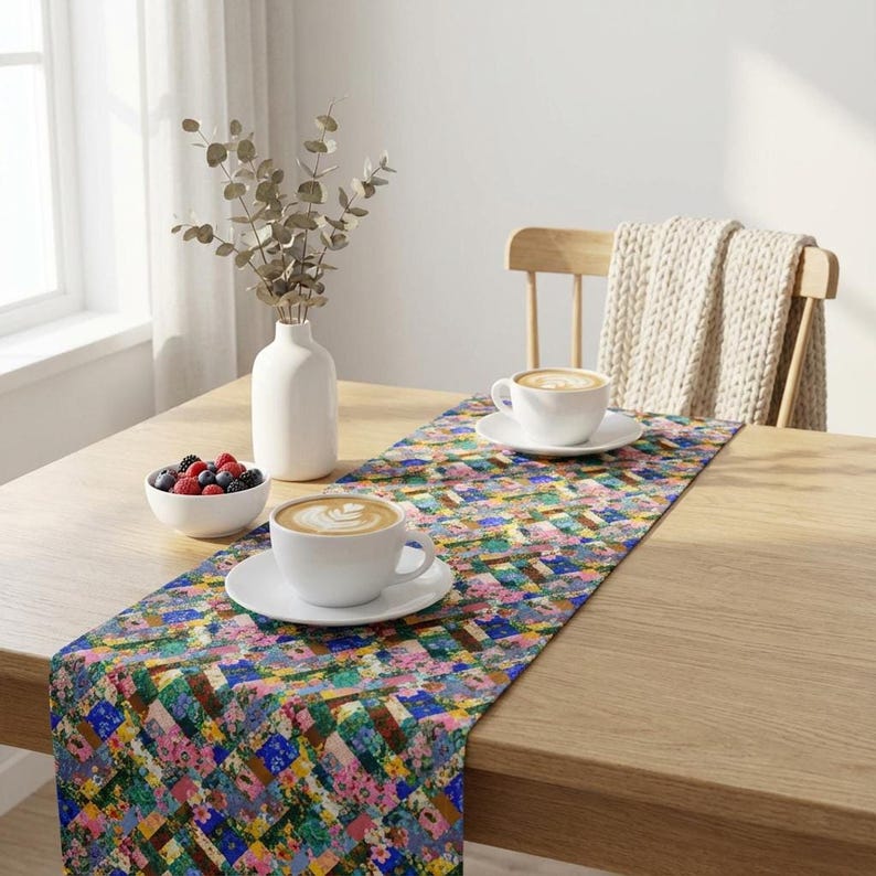 A colorful patchwork floral table runner styled on a light oak wood dining table. The bright morning setup features two large white ceramic mugs with latte coffee foam art, a small white bowl of fresh mixed berries, and dried eucalyptus branches in a modern white ceramic vase by a sunlit window.