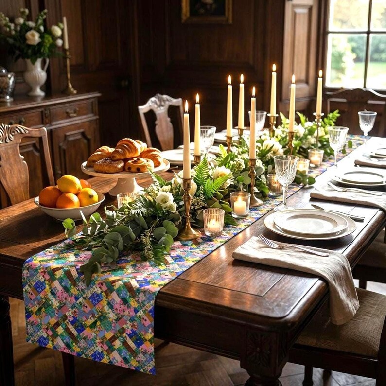 A vibrant patchwork floral table runner styled down a long, formal dark wood banquet table. The elaborate table setting features tall gold taper candlesticks, glowing votive candles, a lush eucalyptus and white flower garland, crystal glassware, a bowl of citrus fruits, and a platter of fresh croissants.