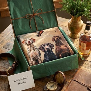 Pode incluir: Um conjunto de presentes com tema de caça, com uma estampa com tema de cães, uma coleira de couro, uma bússola e uma garrafa de uísque escocês. A caixa é forrada com papel de seda verde e amarrada com barbante marrom.