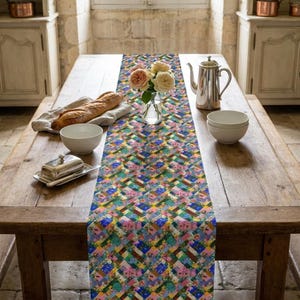 A vibrant patchwork floral table runner styled down a rustic, long wood dining table. The classic French country kitchen setting features a fresh baguette on a linen cloth, a silver tea pot, ceramic bowls, a butter dish, and a glass vase with light pink roses under a bright window.