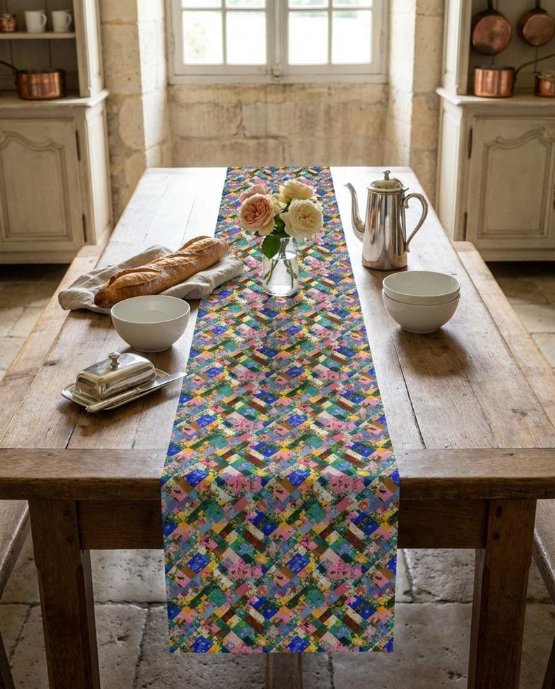 A vibrant patchwork floral table runner styled down a rustic, long wood dining table. The classic French country kitchen setting features a fresh baguette on a linen cloth, a silver tea pot, ceramic bowls, a butter dish, and a glass vase with light pink roses under a bright window.