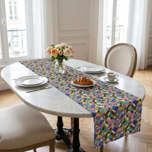A vibrant patchwork floral table runner styled diagonally across an oval white marble dining table. The elegant setup is placed in a bright, sunlit room with chevron wood floors and features a plate with a fresh croissant, white dining plates, a coffee cup, and a glass vase filled with pink and white roses.