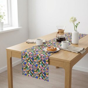 A vibrant patchwork floral table runner styled down a minimalist light oak wood coffee table. The bright dining setup includes two white coffee mugs, a fresh pastry on a ceramic plate, a Chemex glass coffee maker carafe, and a single white flower in a modern white vase against a clean white wall.