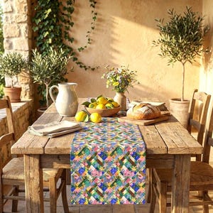 A vibrant patchwork floral table runner styled down a rustic wooden farmhouse table on a sunlit stone patio. The Mediterranean-inspired setup includes a loaf of bread on a cutting board, a wooden bowl filled with fresh lemons, a ceramic pitcher, and a vase of colorful wildflowers, surrounded by potted olive trees.