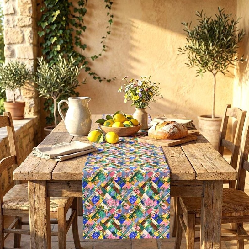 A vibrant patchwork floral table runner styled down a rustic wooden farmhouse table on a sunlit stone patio. The Mediterranean-inspired setup includes a loaf of bread on a cutting board, a wooden bowl filled with fresh lemons, a ceramic pitcher, and a vase of colorful wildflowers, surrounded by potted olive trees.