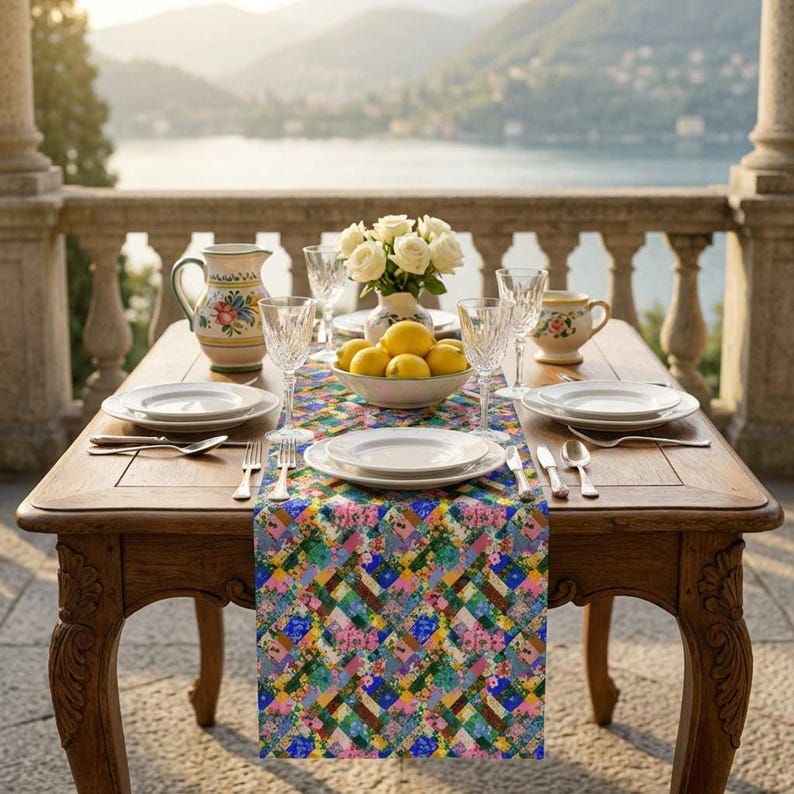 A colorful patchwork floral table runner styled on an ornate wooden dining table on a luxury balcony overlooking a lake. The table setup features white plates, crystal glasses, a bowl of fresh lemons, white roses, and Italian ceramic pitchers against a scenic mountain and water backdrop.