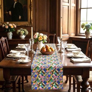 A vibrant patchwork floral table runner styled down a formal dark wood dining table. The classic interior features ornate wood paneling, fine china tableware, crystal goblets, silver candlesticks, and a center wooden bowl filled with fresh pears. Elegant roses and oil paintings decorate the background.